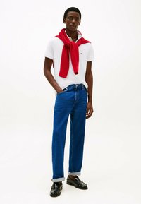Witte polo met korte mouwen, met een rode trui om de nek, blauwe spijkerbroek en zwarte loafers met een glanzende afwerking.