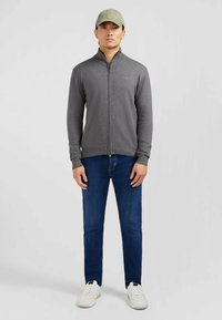 Veste zippée grise avec col montant, associée à un jean bleu et des baskets blanches. Le modèle porte une casquette vert clair. Design simple, style ajusté.