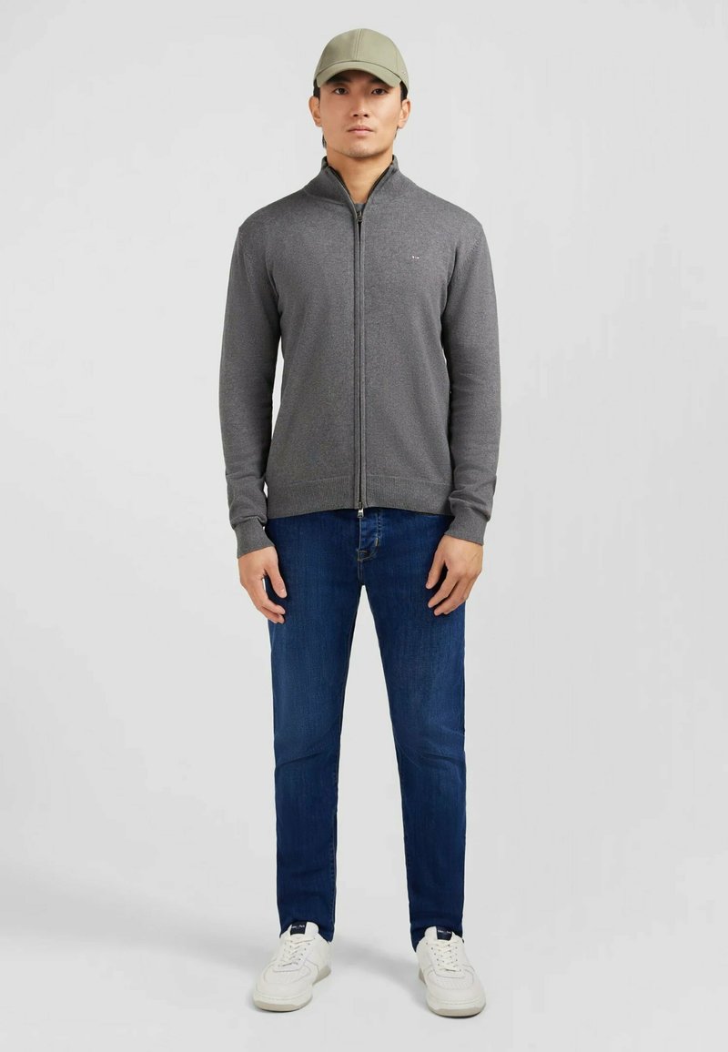 Veste zippée grise avec col montant, associée à un jean bleu et des baskets blanches. Le modèle porte une casquette vert clair. Design simple, style ajusté.