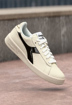 Cream leather sneaker with a black patent accent, round toe, flat white laces, and textured rubber sole featuring a logo on the tongue.
