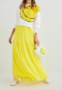 Bílá mikina s žlutým smajlíkem, doplněná průsvitnou žlutou maxi sukní a malou bílou kabelkou s motivem smajlíka.