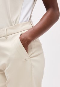 Σκούρο δέρμα στο χέρι με το χέρι μισοκρυμμένο στην τσέπη παντελονιού ψηλόμεσου σε εκρού χρώμα, μπροστά σε απλό φόντο.