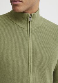 Strikket zip-genser i lys grønn, med høy krage og lett teksturert stoff med en glatt glidelås foran.