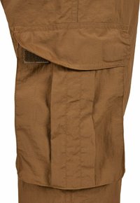 Bruine cargobroek met een grote zijzak met klittenbandsluiting, gemaakt van lichte stof met een subtiele textuur en stikdetails.
