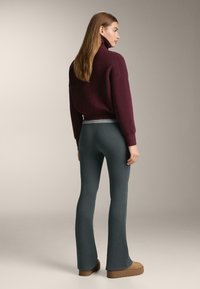 Vínová svetr s vysokým límcem a žebrovanou texturou, spárovaný s tyrkysovými žebrovanými rozšířenými kalhotami a světle hnědými platformovými botami. Jednoduché pozadí.