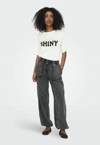 Off-white kortærmet T-shirt med "SHINY" tekst og sorte accenter. Løse grå denimbukser med elastisk talje og sidelommer. Sorte prikkede flade sko.