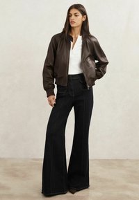 Jachetă bomber din piele maro cu manșete plisate, asortată cu blugi din denim întunecat, cu talie înaltă și cusături galbene contrastante, pe un fundal neutru.