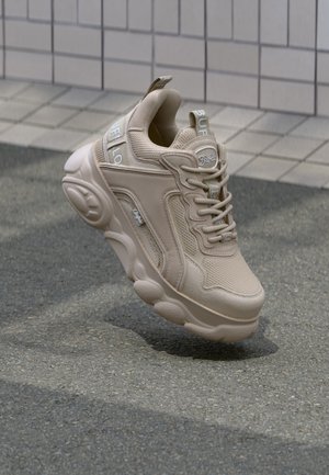 Beige chunky sneaker met "Buffalo"-branding, gestructureerd bovenwerk, dikke zool en veters, geplaatst boven een geplaveide grond en een achtergrond van betegelde muur.