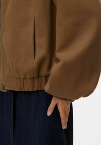 Veste polaire marron avec fermeture éclair à l'avant, taille élastiquée et manches bouffantes. Associée à une jupe en jean foncé.