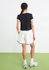 Joven con el cabello trenzado, vistiendo una camiseta negra con ribetes blancos, pantalones cortos deportivos blancos con ribetes en azul marino, calcetines blancos y zapatillas plateadas.