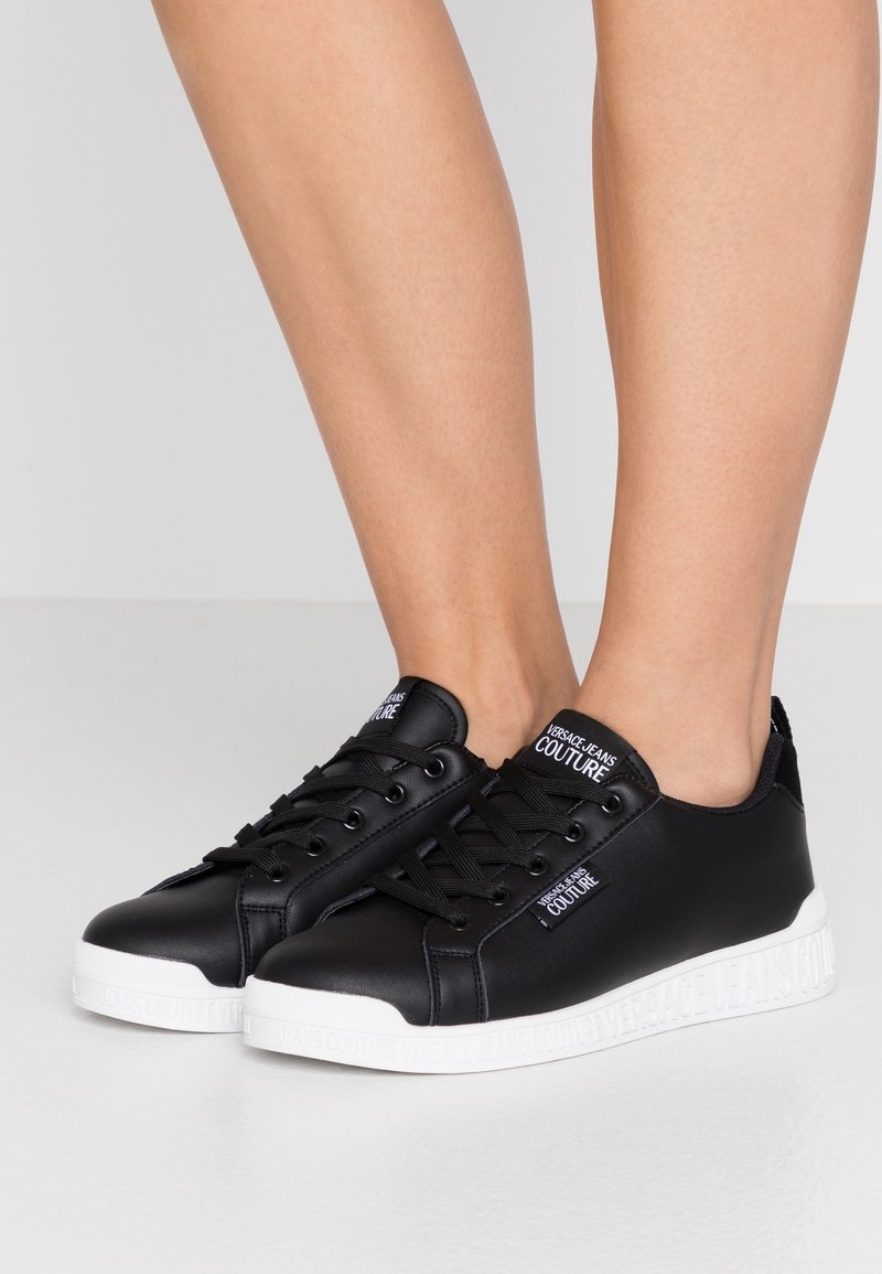 Černé syntetické tenisky s hladkým povrchem, bílou gumovou podrážkou a označením značky na jazyku a na boku. Šněrovací design s kulatou špičkou.