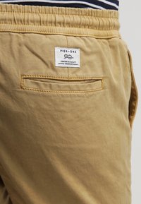 Beige avslappnade byxor med elastisk midja och bakfickor, med en Pier One etikett för komfort och kvalitet.