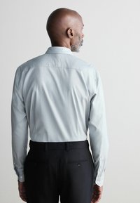 Light grey patterned long-sleeve shirt with buttoned cuffs, paired with black trousers featuring back pocket accents.