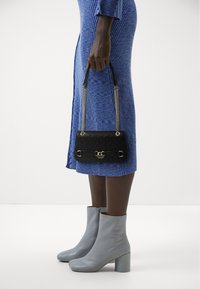 Borsa a tracolla nera trapuntata con hardware dorato e manico a catena, tenuta in mano. Indossata con un vestito blu a coste e stivaletti grigi con tacco.