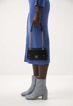 Borsa a tracolla nera trapuntata con hardware dorato e manico a catena, tenuta in mano. Indossata con un vestito blu a coste e stivaletti grigi con tacco.