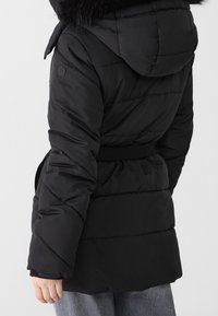 Person wearing a black quilted puffer jacket with a hood trimmed in black fur and a cinched waist, paired with gray pants.