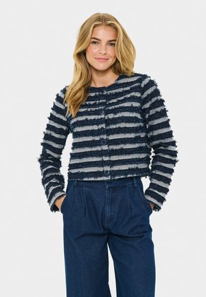Vrouw met haar handen in haar zakken, gekleed in een marineblauw en grijs gestreept knooptrui met structuur en een hoge taille donkerblauwe broek op een witte achtergrond.