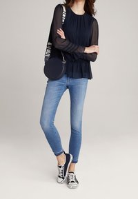 Marinebluse mit transparenten Ärmeln, Rüschensaum, kombiniert mit hellblauen Skinny-Jeans und marineblauen Sneakers mit weißen Akzenten und Logo.