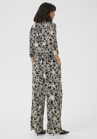 Floral print jumpsuit in black and cream, featuring three-quarter sleeves, pockets, a loose fit, and a tied waist detail.