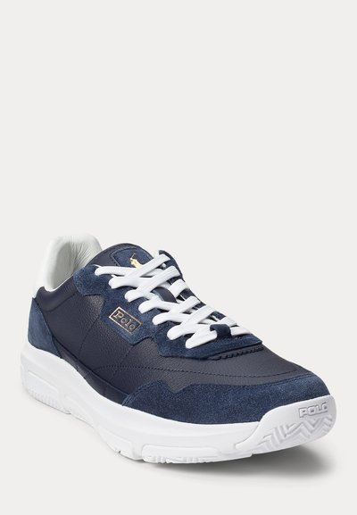 Tengerészeti bőr és velúr sportcipő, fehér fűzőkkel és talppal, arany logóval az oldalon és texturált részletezéssel.