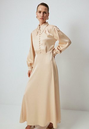 Mujer con un vestido largo de satén beige con mangas abullonadas, botones forrados de tela y un cuello ancho, de pie con una mano en la cadera.