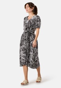 Schwarz-weiß gestreiftes Midi-Kleid im Zebra-Stil mit Puffärmeln, V-Ausschnitt und gebundenem Taillengürtel. Modell trägt beige strukturierte Sandalen.