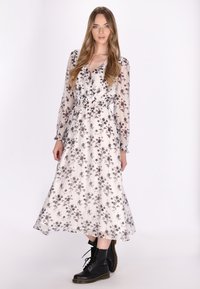 Bloemrijke maxi-jurk met lange mouwen, wit en grijs patroon, aangesloten taille, gecombineerd met zwarte veterschoenen, gladde stoftextuur.