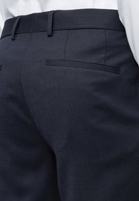 Κοντινό πλάνο ναυτικού μπλε ραμμένων παντελονιών με θηλιές ζώνης και πίσω τσέπες welt, φορεμένα με λευκό πουκάμισο που είναι περασμένο μέσα.