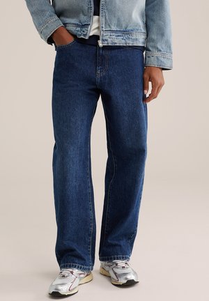 Homme portant un jean bleu foncé ample, une veste en jean bleu clair et des baskets blanches, debout avec une main dans la poche.