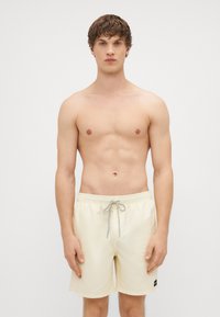 Bañador amarillo claro con cinturilla elástica y cordón gris, que cuenta con bolsillos laterales y una pequeña etiqueta de marca negra.