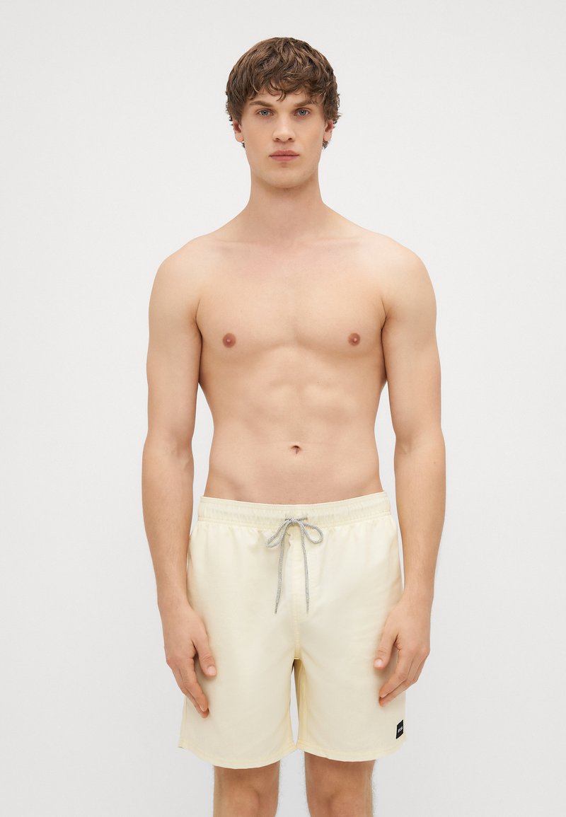 Calções de banho amarelo-claro com cintura elástica e cordão cinza, apresentando bolsos laterais e uma pequena etiqueta de marca preta.