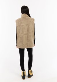 Gilet senza maniche in pelliccia sintetica color miele con colletto alto e dettagli cuciti, indossato sopra un top a maniche lunghe nero e abbinato a leggings neri e stivali.