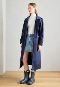 Giacca trench navy, lunga con bottoni e bavero; maglione a collo alto bianco, gonna di jeans; stivali blu impermeabili con manici e lati texture.