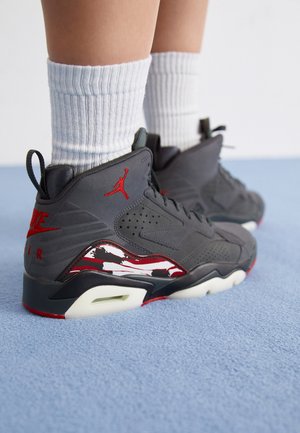 Osoba nosząca szare i czerwone trampki Air Jordan z białymi skarpetkami, stojąca na jasnoniebieskim dywanie.