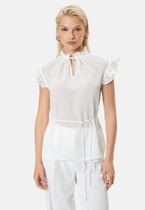 Blonde vrouw die een witte mouwloze blouse met ruches en een strik bij de hals draagt, samen met een bijpassende witte broek, staand tegen een effen achtergrond.