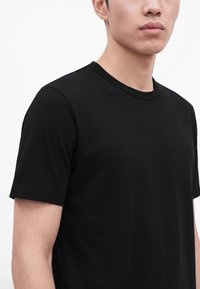 Άνδρας που φοράει απλό μαύρο κοντομάνικο T-shirt, εμφανιζόμενος από τους ώμους μέχρι το πηγούνι, σε λευκό φόντο.