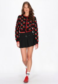 Svart stickad cardigan med mönster av röda körsbär, röda detaljer och knappar, i kombination med en svart plisserad kjol och röda sneakers.