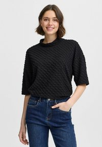 Schwarzes, strukturiertes Oberteil mit hohem Kragen, kurzen Ärmeln und einem gemusterten Oberfläche. Kombiniert mit dunkelblauen Jeans.
