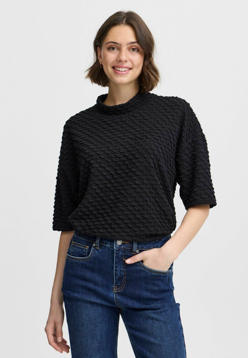 Schwarzes, strukturiertes Oberteil mit hohem Kragen, kurzen Ärmeln und einem gemusterten Oberfläche. Kombiniert mit dunkelblauen Jeans.