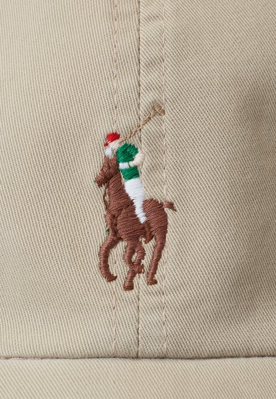 Bēšas krāsas auduma cepure ar brūnu izšūtu polo spēlētāju uz zirga, kuram ir sarkans un zaļš braucēja tērps un balta zirdziņa sedziņa.