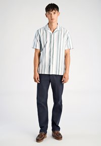 Jonge man die staat, gekleed in een wit en grijs verticaal gestreept kortemouwen shirt, donkerblauwe broek en bruine veterschoenen.