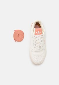 Zapatilla deportiva blanca con detalles perforados y acentos en naranja claro. Parte superior de cuero liso, cordones planos blancos y cordones redondos en naranja claro.