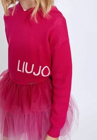Fuchsia huvtröja med "LIUJO" i vit text, tillsammans med en lager av tyllkjol i en ljusare rosa nyans. Mjuk stickad tyg med ribbade ärmslut.
