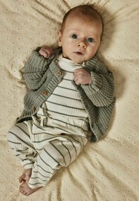 Grå stickad kofta med träknappar, lager ovanpå en vit randig bodysuit och byxor, vilande på en texturerad, ljus färgbakgrund.