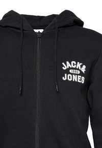 Sort zip-up hættetrøje lavet af blødt stof; har snørre hætte og hvid "JACK & JONES 1990" logo på brystet.