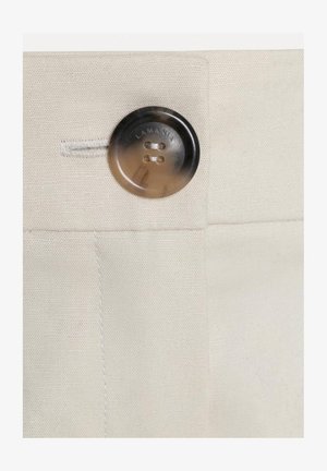 Tumepruuni ümmargune nupp, millel on neli auku ja mis on kinnitatud beežile kangale, kus lähedal on õmmeldud nööbiaugu.