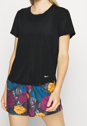 Femme portant un t-shirt Nike noir perforé et un short à fleurs avec des fleurs bleues, violettes, jaunes et grises sur un fond sombre.