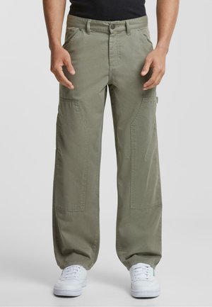 Pantalones cargo verde oliva hechos de tela, con un corte holgado, grandes bolsillos laterales y cierre de cremallera, combinados con zapatillas blancas.