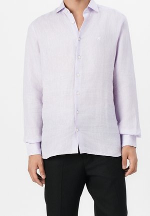 Homme portant une chemise lavande claire à manches longues boutonnée avec un pantalon noir, debout devant un fond blanc uni.