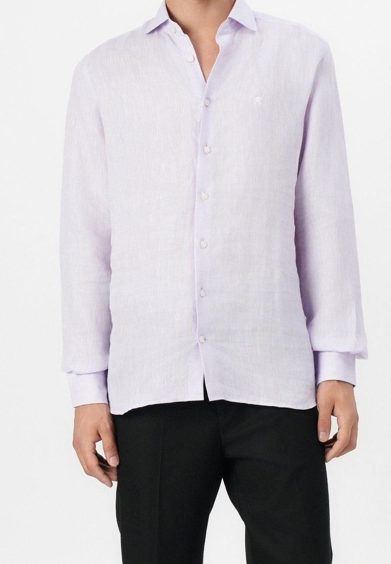 Homme portant une chemise lavande claire à manches longues boutonnée avec un pantalon noir, debout devant un fond blanc uni.